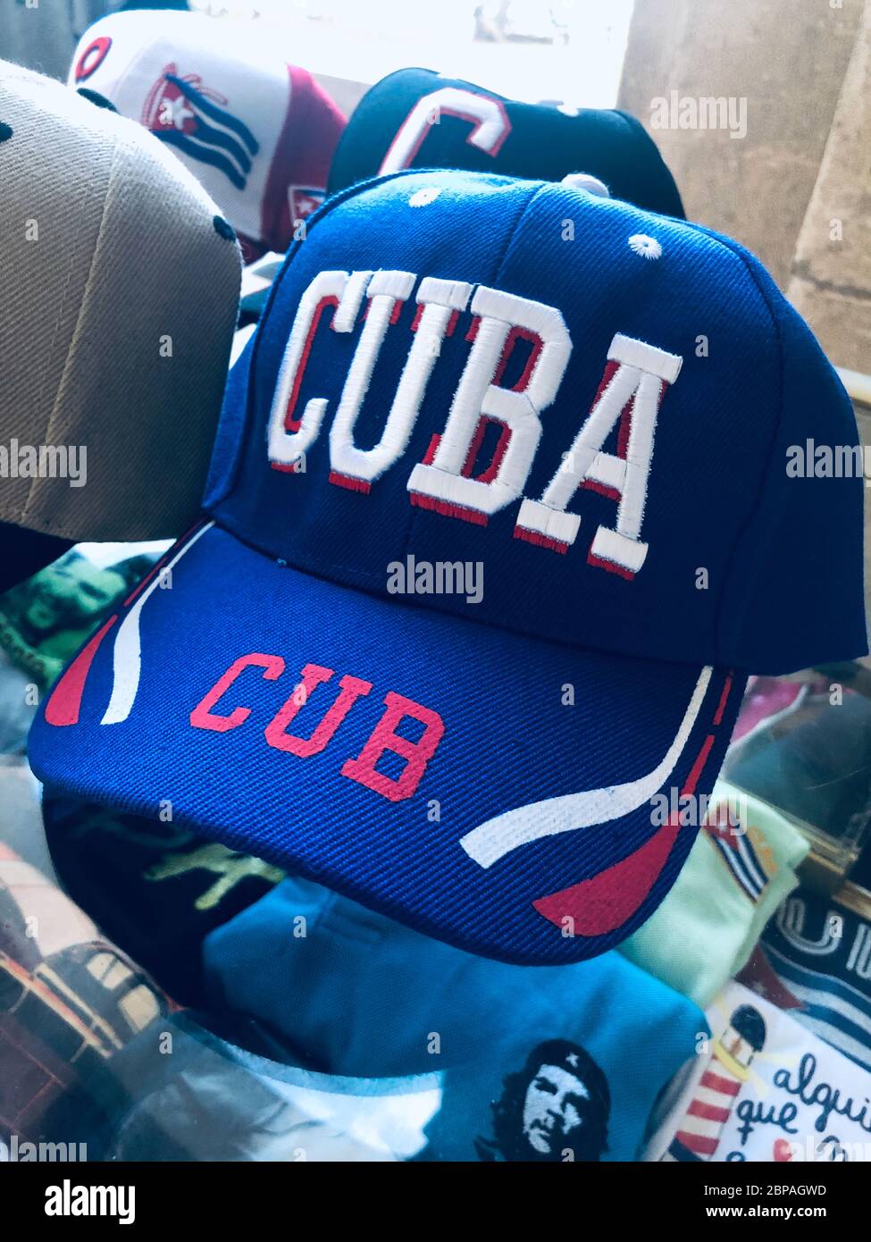 Imagen de una gorra de béisbol azul roja y blanca con Cuba escrita en letras  grandes. Sentado en la parte superior de una caja con otras gorras, y  camisetas con la cara