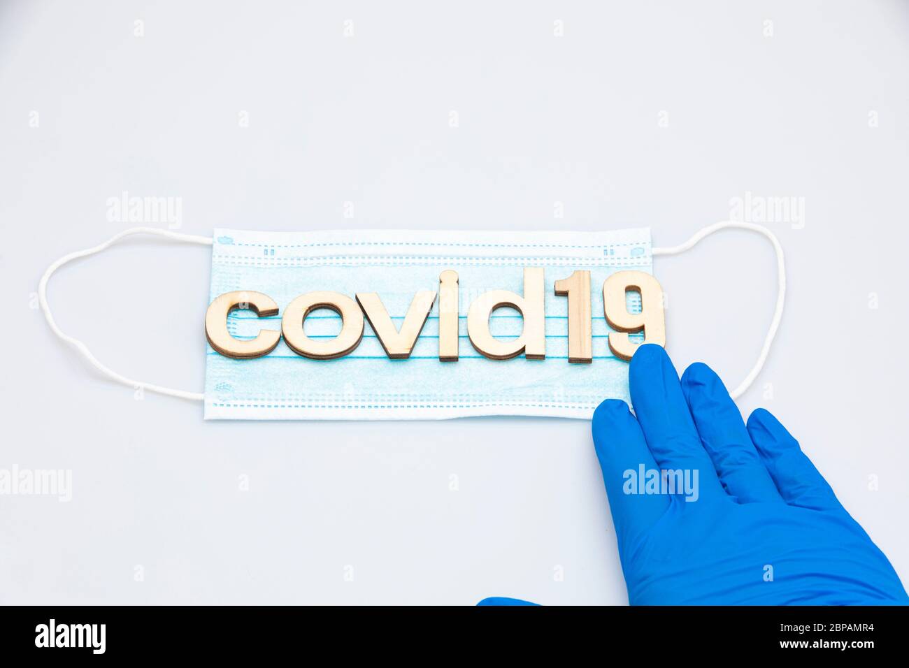mano con un guante quirúrgico azul que sostiene la última parte de la palabra covid19 formado con letras colocadas sobre una máscara quirúrgica aislada sobre un fondo blanco Foto de stock