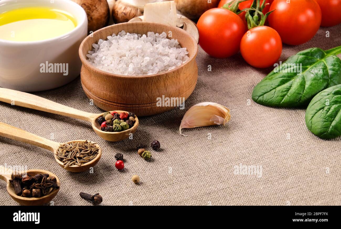 Diversas especias en pequeñas cucharas de madera, sal, aceite vegetal, tomates cherry, champiñones, espinacas y ajo en un tejido de lino. Foto de stock