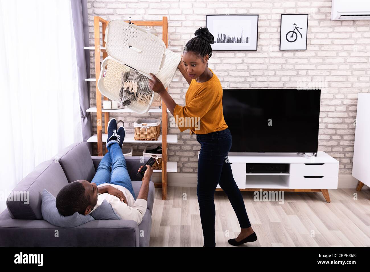 Mujer joven arrojando la ropa sucia en perezoso marido tumbado en el sofá Foto de stock