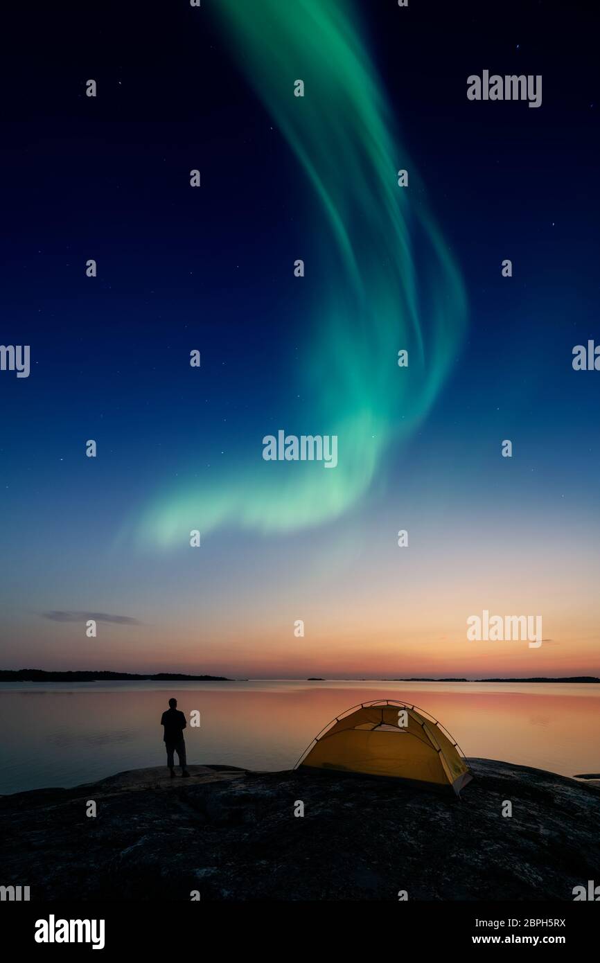 Hombre mirando las luces del norte alias Aurora Borealis junto a una tienda de campaña un mar tranquilo. Foto de stock