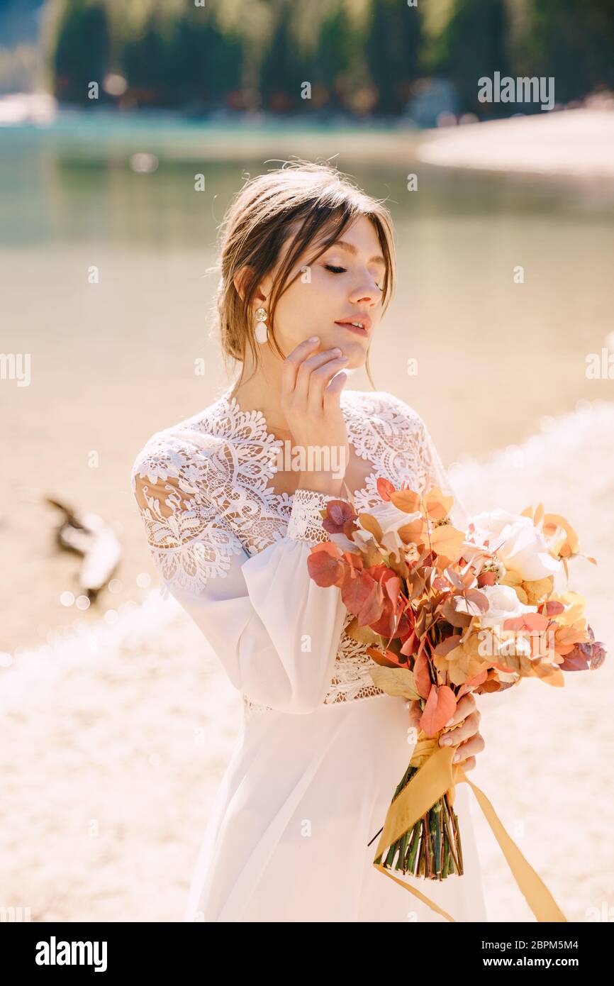 Hermosa novia en un vestido blanco con mangas y encaje, con un ramo de  otoño amarillo de flores secas y rosas de peony, en el Lago di Braies en  Italia Fotografía de