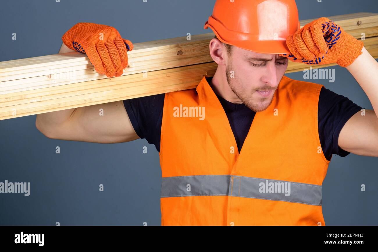 Carpenter, trabajador de madera, fuerte constructor en cara ocupada lleva  viga de madera en el hombro. El hombre en guantes protectores sostiene  visera de casco protector, fondo gris. Concepto de seguridad y