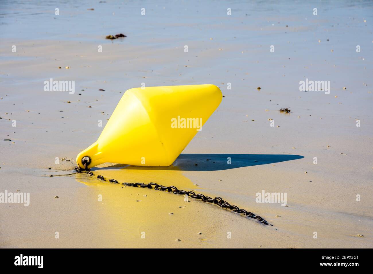 Una boya amarilla grande y su cadena de anclaje, utilizada como marcador de canal de lanzamiento, tumbado en la arena húmeda a marea baja en la playa al sol. Foto de stock