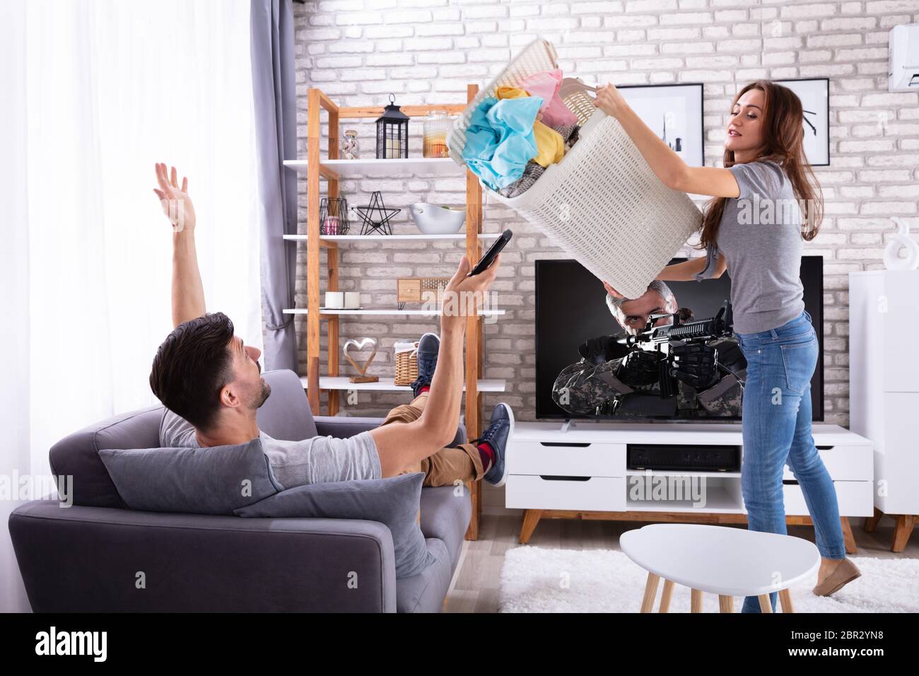 Mujer joven arrojando la ropa sucia en perezoso marido tumbado en el sofá Foto de stock