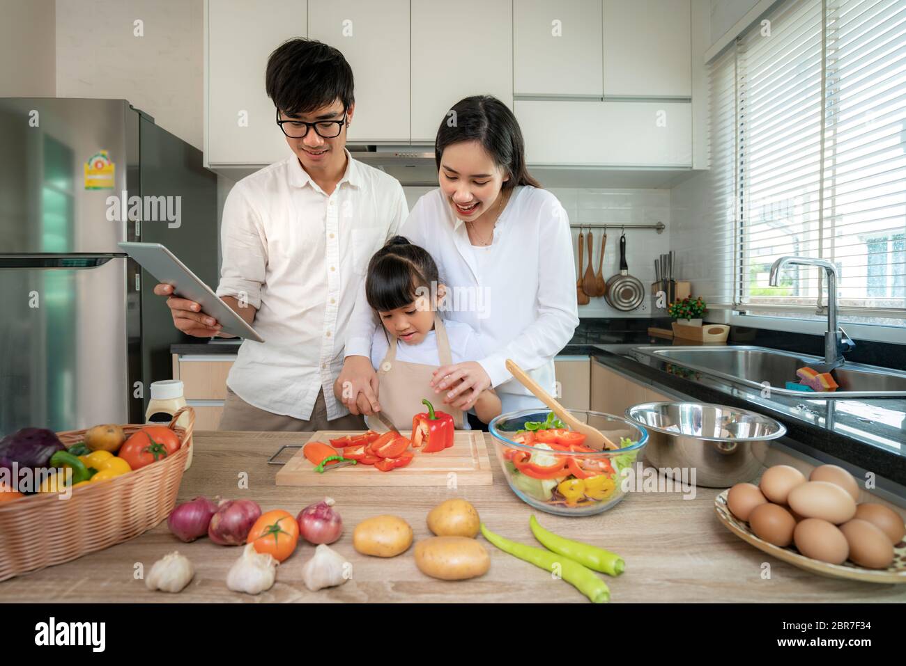 Madre asiática que enseña cortar verduras a las hijas y padre de pie cuando una familia cocina en la cocina en casa. Vida familiar amor relación, Foto de stock