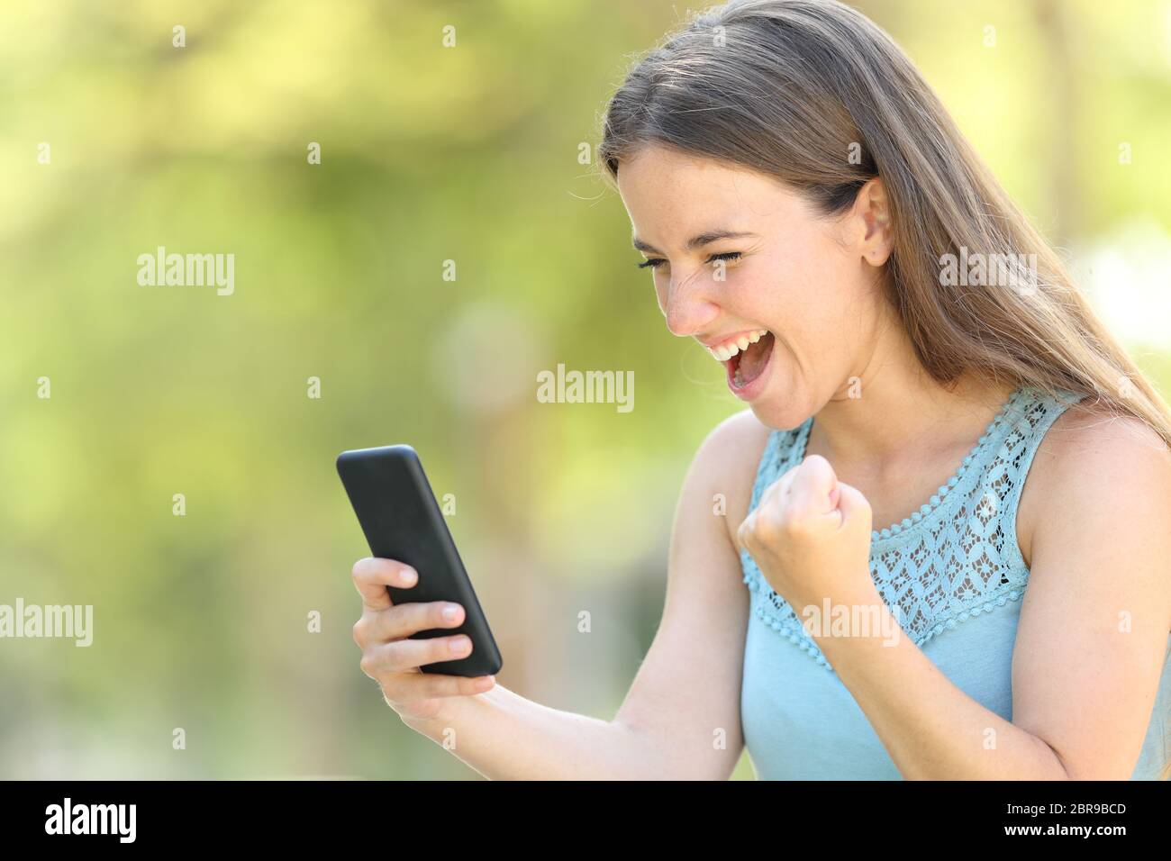 Chica encontrar interesantes contenidos en línea en el teléfono inteligente en un parque con un fondo verde Foto de stock