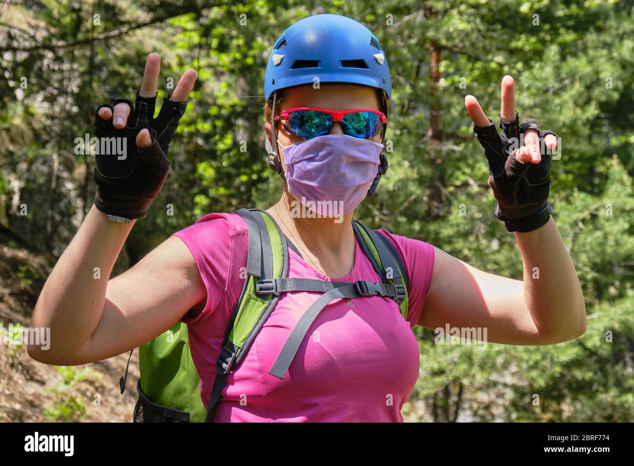Mujer alpinista con una máscara médica y casco, muestra un signo de victoria con ambas manos. Concepto de Coronavirus (Covid-19) restricciones que se están aliviando y. Foto de stock