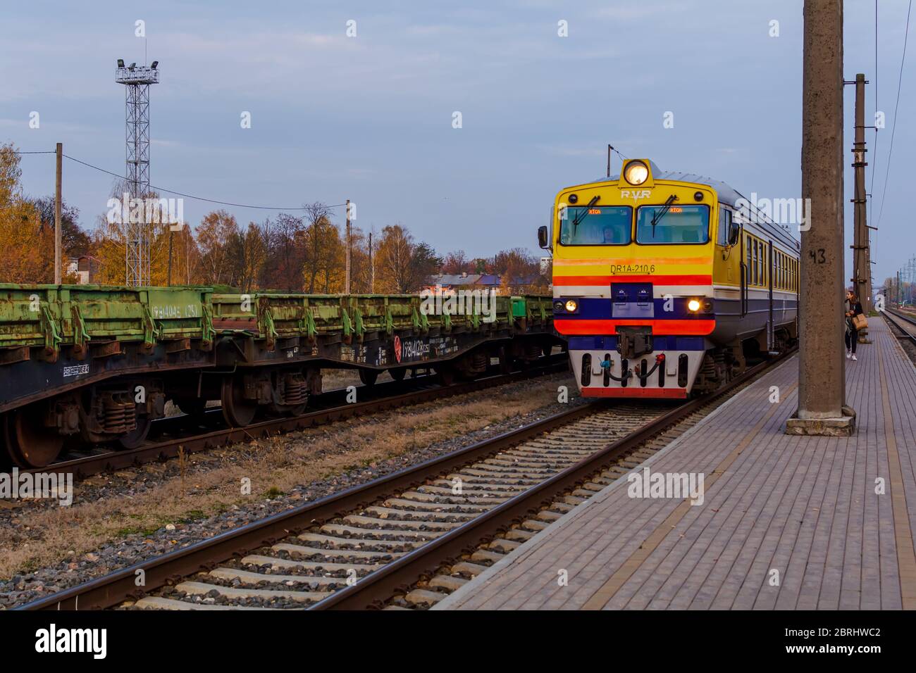 Tren suburbano de pasajeros que llega a la plataforma de la estación por la noche. Frente amarillo-azul del tren en la estación de tren de Riga. Transporte CO Foto de stock