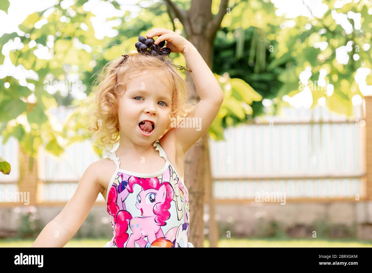Odessa - 25 08 2019 niña pequeña jugar lengua con uva usando traje de baño con estampado de poni. Rubia caucásica emocional feliz alegría Fotografía de stock - Alamy