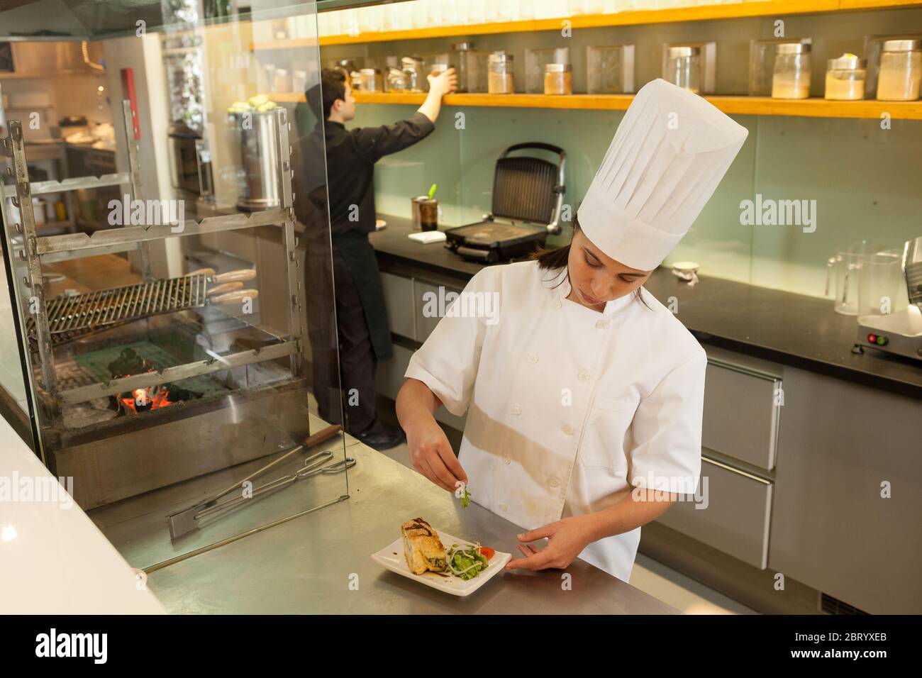 Chef mujer con sombrero de chef de pie en la encimera en la cocina comercial, guarnición plato de comida. Foto de stock