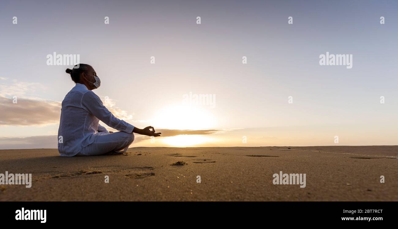 hombre meditando en máscara médica en lotus pose en la playa, hombre usando máscara quirúrgica y ropa de yoga blanca con peinado de nudo superior al amanecer Foto de stock