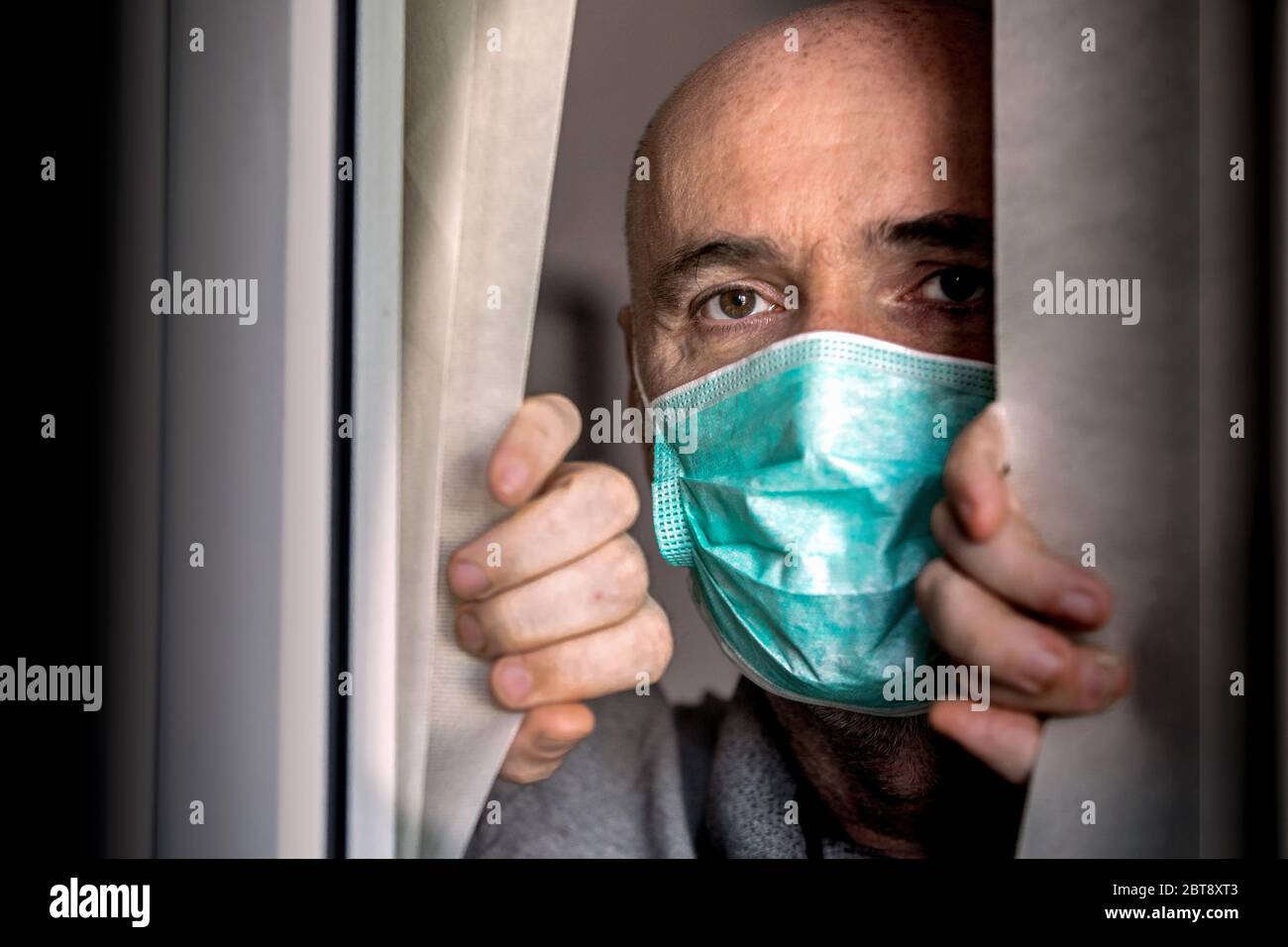 El hombre que lleva una máscara médica mirando a través de la ventana. Aislamiento en casa para el brote de virus. Concepto de autocuarentena en el hogar, prevención COVID-19. Foto de stock