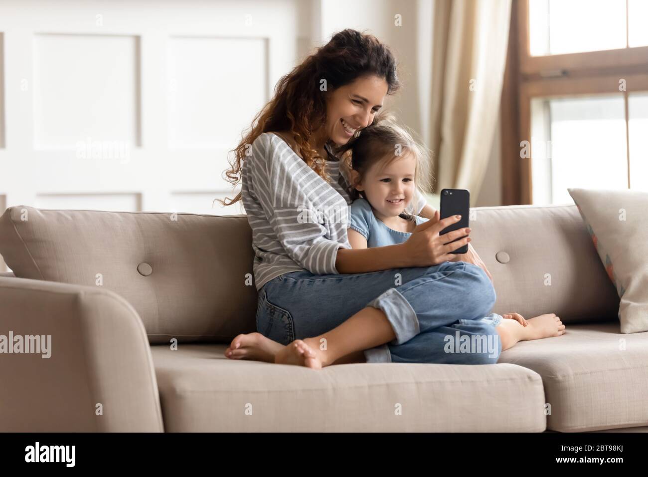 Sonriendo madre y pequeña hija abrazando, usando el teléfono juntos Foto de stock