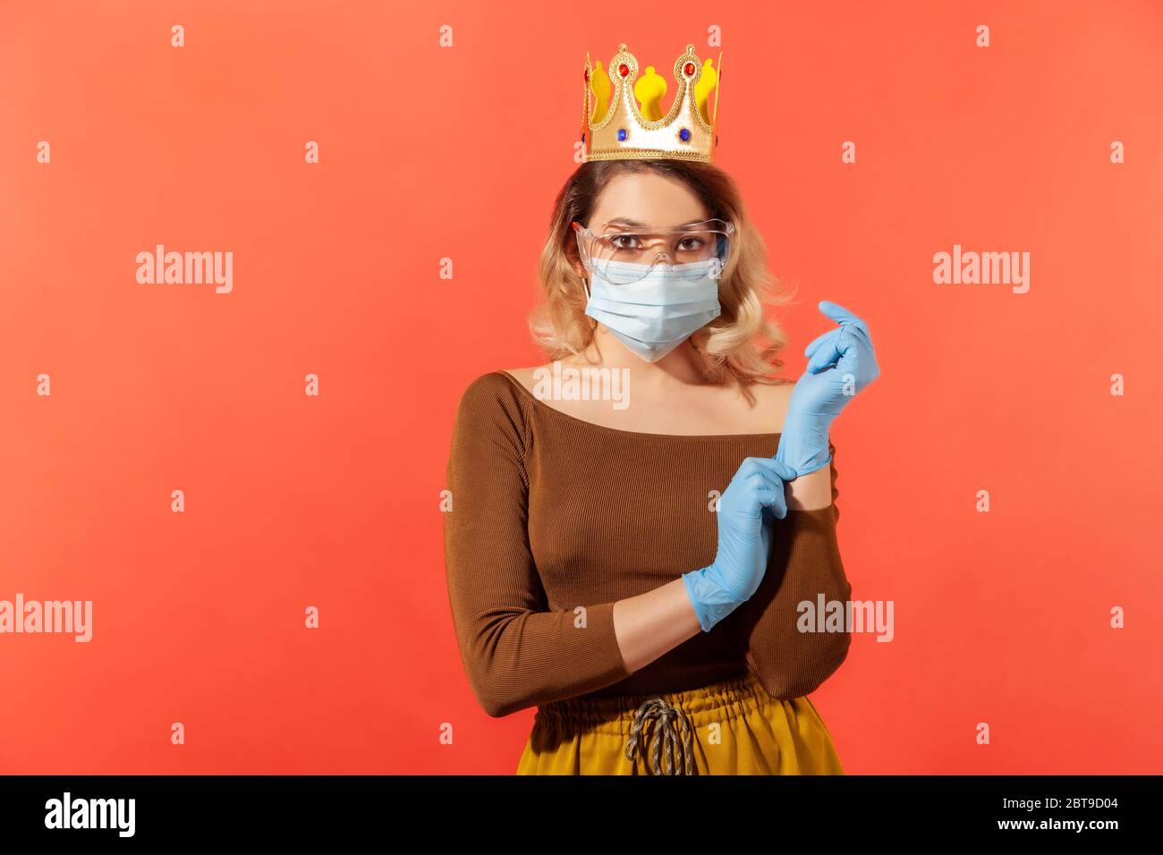 Mujer con corona en la cabeza, en máscara higiénica y gafas protectoras, ajustando guantes de seguridad para prevenir el coronavirus contagioso de la gripe, enfermedad de 2019 ncov. i Foto de stock
