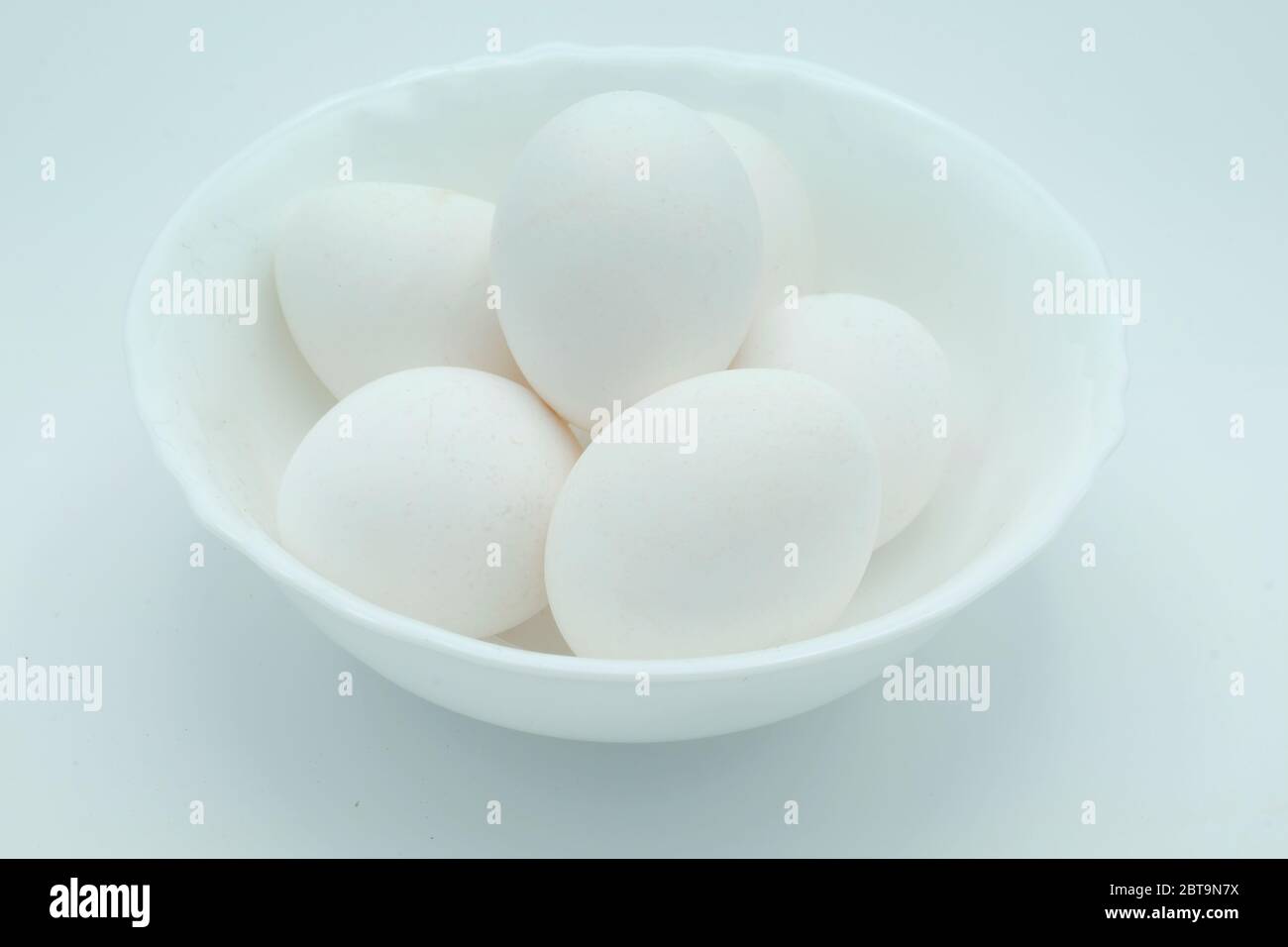 Un tazón blanco lleno de huevos blancos de gama libre sobre un fondo blanco Foto de stock