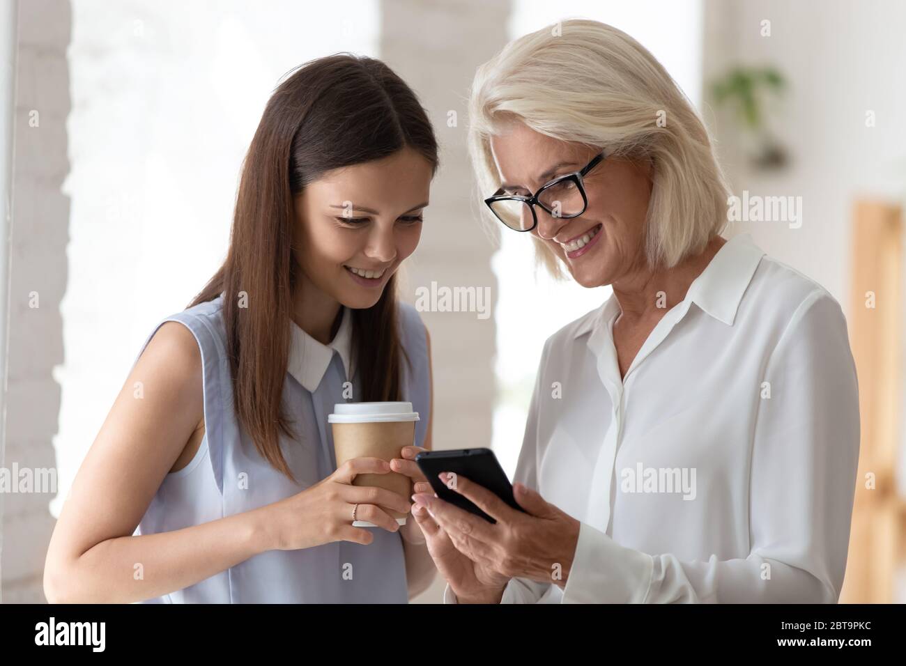 Las mujeres sonrientes hojean el smartphone en la cafetería de la oficina Foto de stock