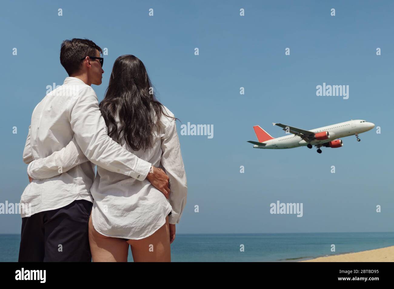 novia en bikini y novio en camisa blanca se pone de pie junto al océano  inmenso y mira el avión bajo el cielo azul vista de la parte trasera  Fotografía de stock -
