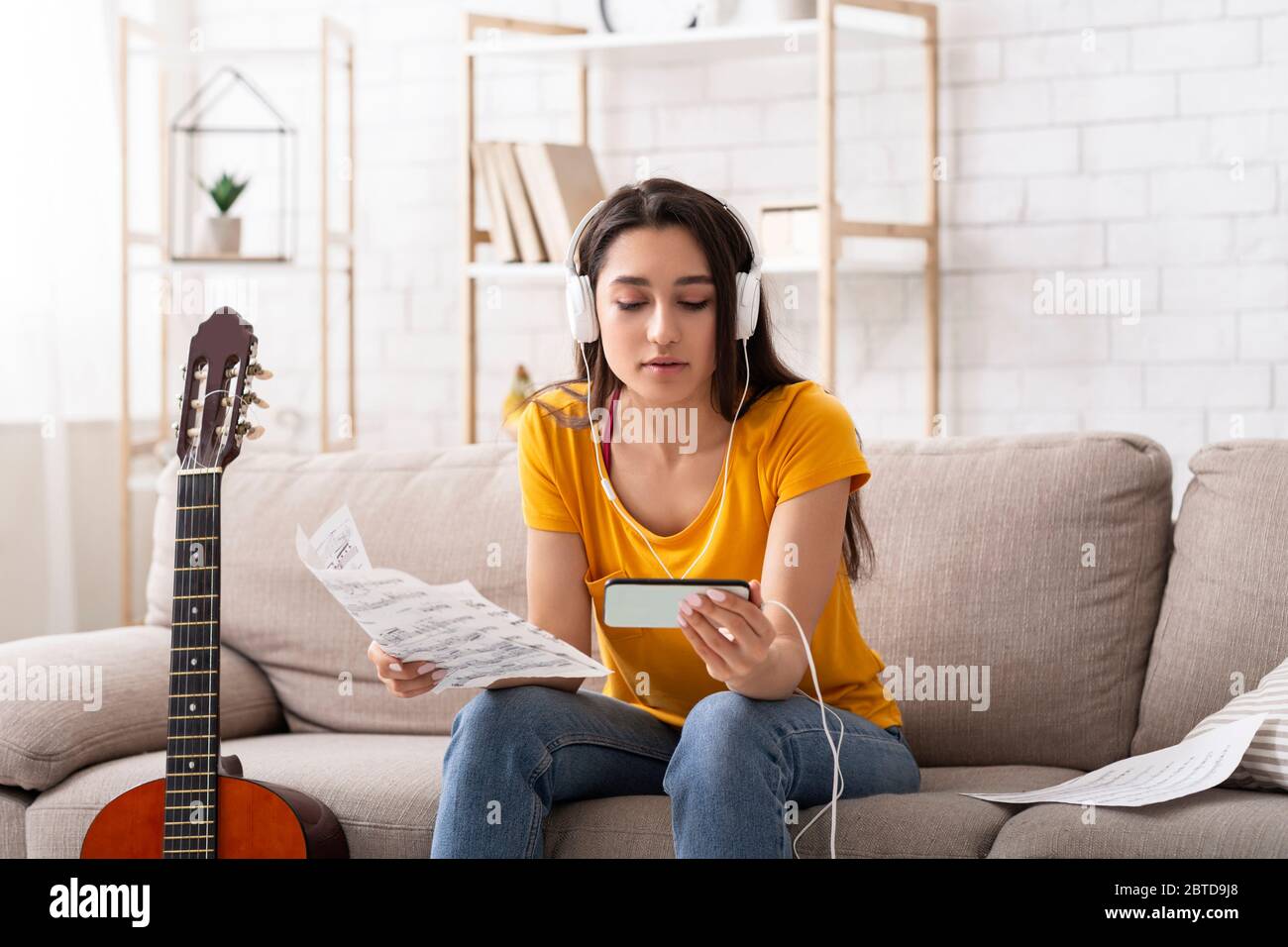 Milenial niña en auriculares aprendiendo a tocar la guitarra viendo video  tutorial en línea en el teléfono móvil, en interiores Fotografía de stock -  Alamy