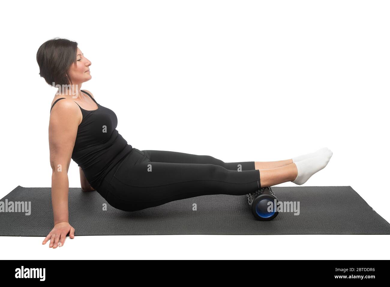 Una mujer de mediana edad en una esterilla gimnástica con rodillo miofascial hace un ejercicio en sus caderas sobre un fondo blanco. Foto de stock