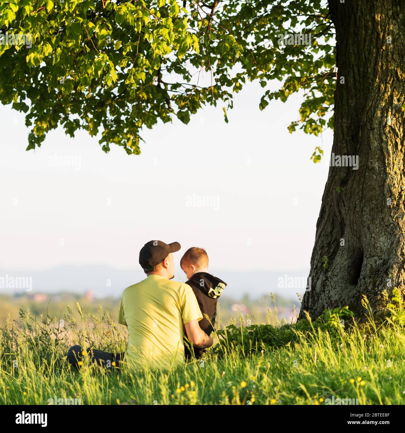 Papá con hijo en el prado de primavera sentado bajo el árbol en hierba alta. Concepto de viaje con niños Foto de stock