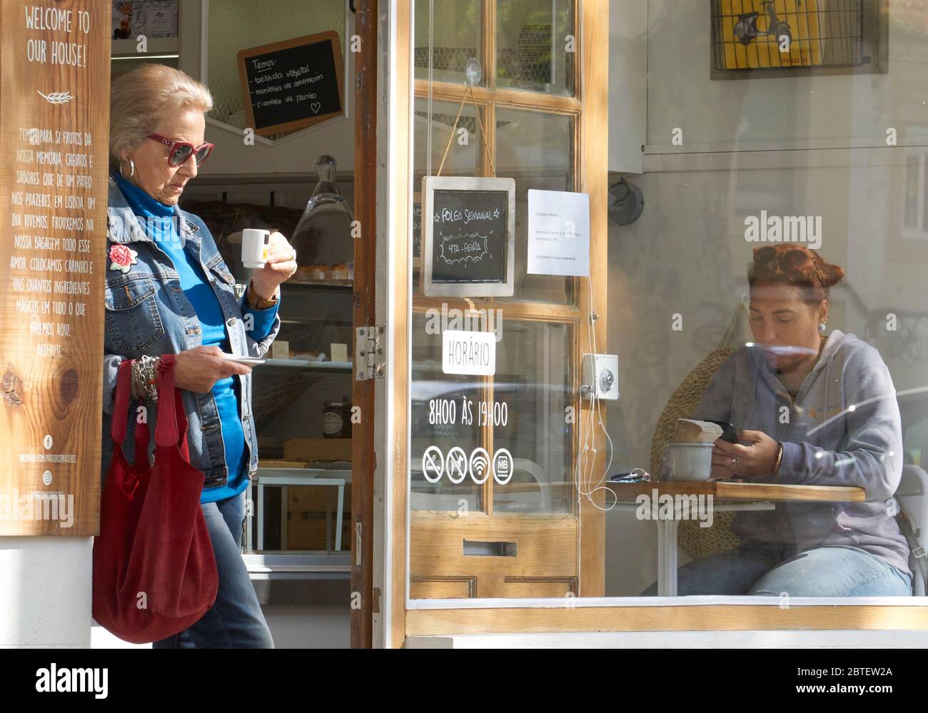 Una mujer mayor tomando café de pie en la entrada de un café ordenado en Ericeira, Portugal, una mujer joven sentada en una mesa en una ventana grande Foto de stock