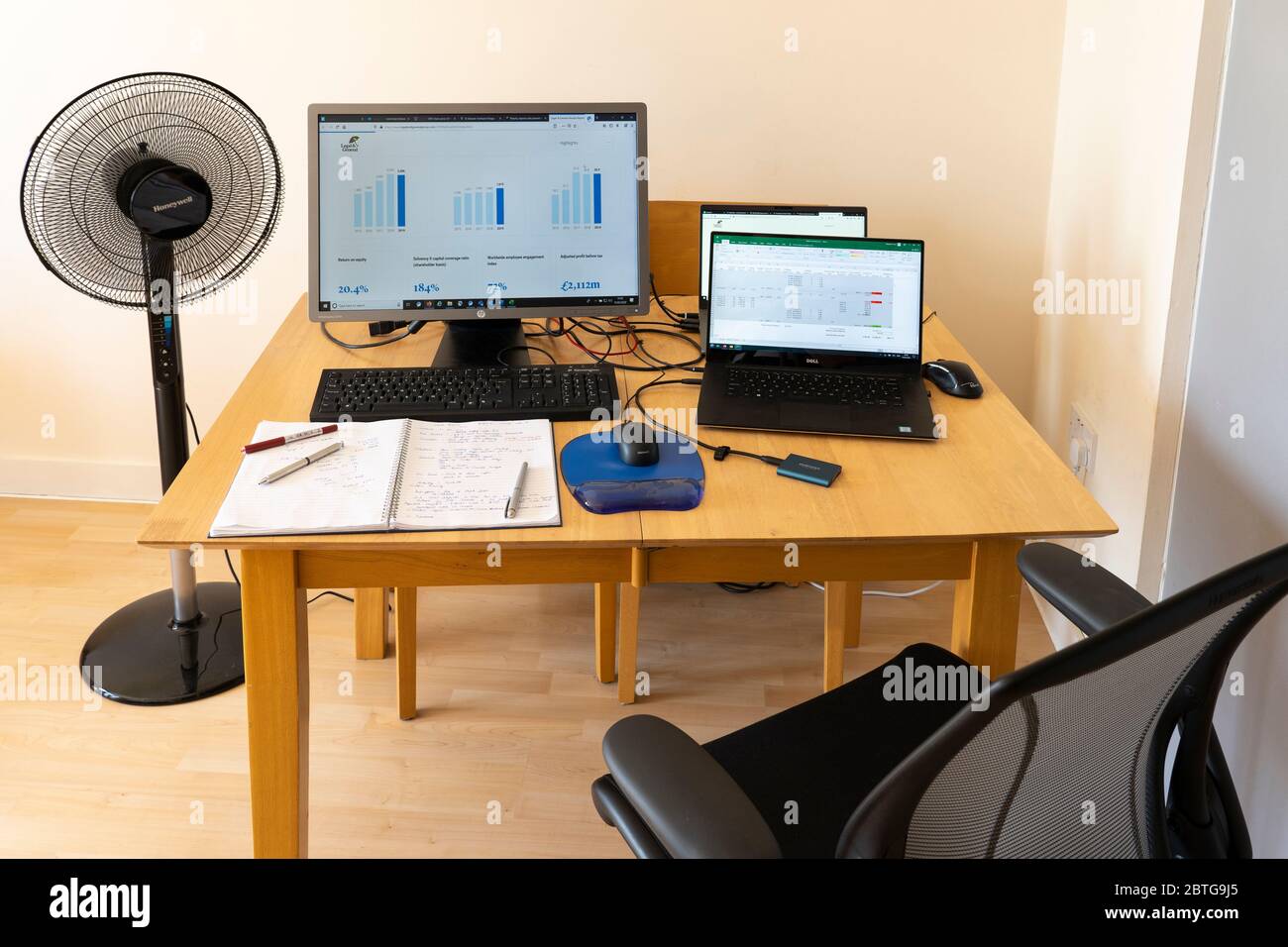 Una mesa con ordenadores portátiles y un monitor con una alfombrilla para ratón y ratón, bloc de notas y bolígrafos, un ventilador y pantallas que muestran gráficos. Tema - trabajando desde casa. REINO UNIDO Foto de stock