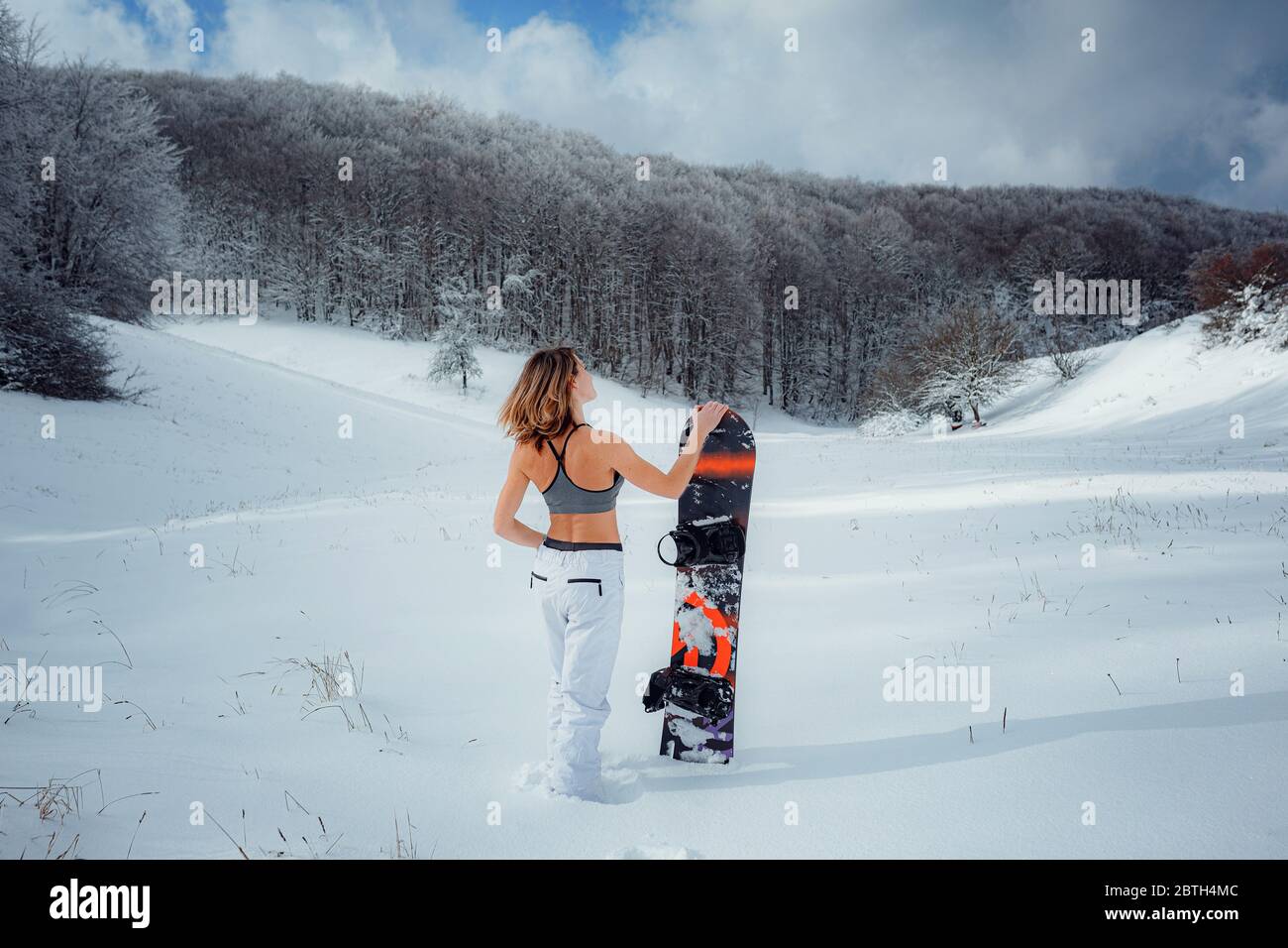 Snowboarder femenina sostiene snowboard e va a snowboard. Actividad deportiva de invierno, nieve en el bosque estilo de vida al aire libre. Chica con una camiseta corta y esquí w Foto de stock