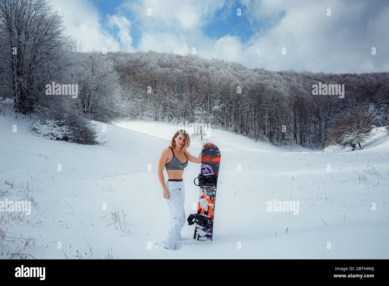 Snowboarder femenina sostiene snowboard e va a snowboard. Actividad deportiva de invierno, nieve en el bosque estilo de vida al aire libre. Chica con una camiseta corta y esquí w Foto de stock