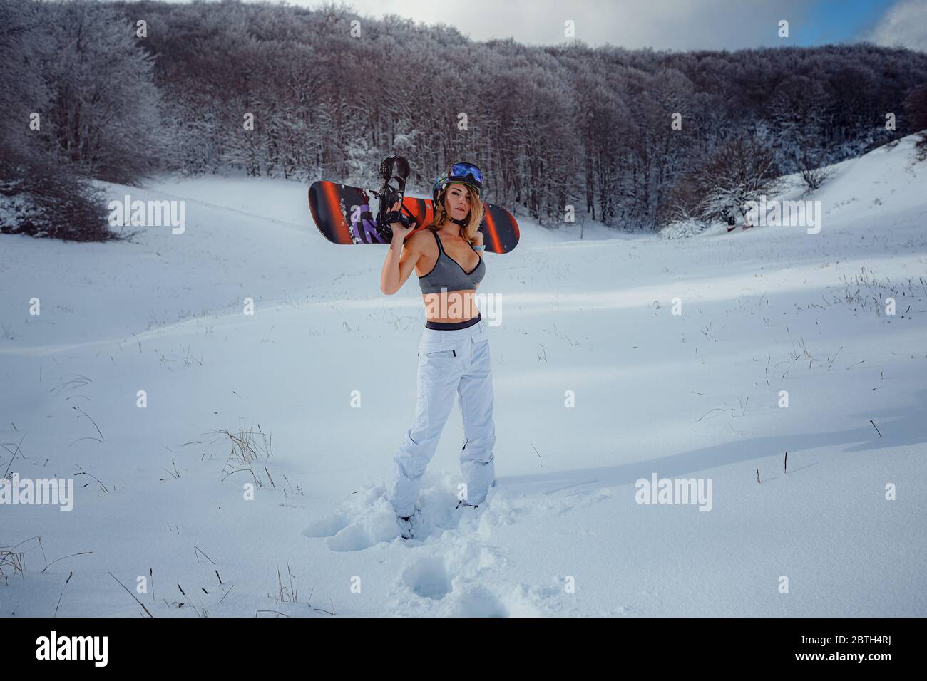 Snowboarder femenina sostiene snowboard e va a snowboard. Actividad deportiva de invierno, nieve en el bosque estilo de vida al aire libre. Chica con una camiseta corta y esquí w Foto de stock