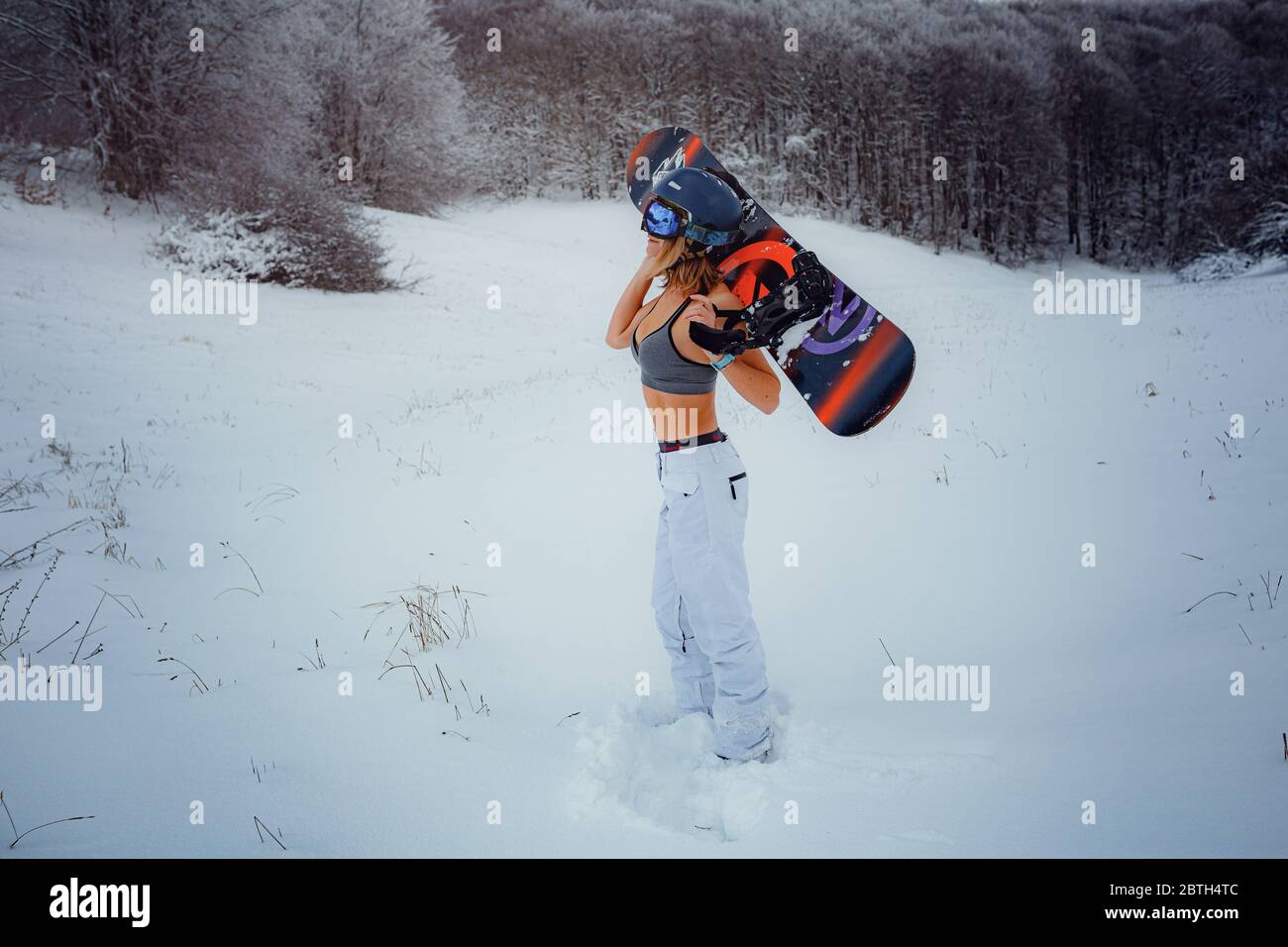 Snowboarder femenina sostiene snowboard e va a snowboard. Actividad deportiva de invierno, nieve en el bosque estilo de vida al aire libre. Chica con una camiseta corta y esquí w Foto de stock