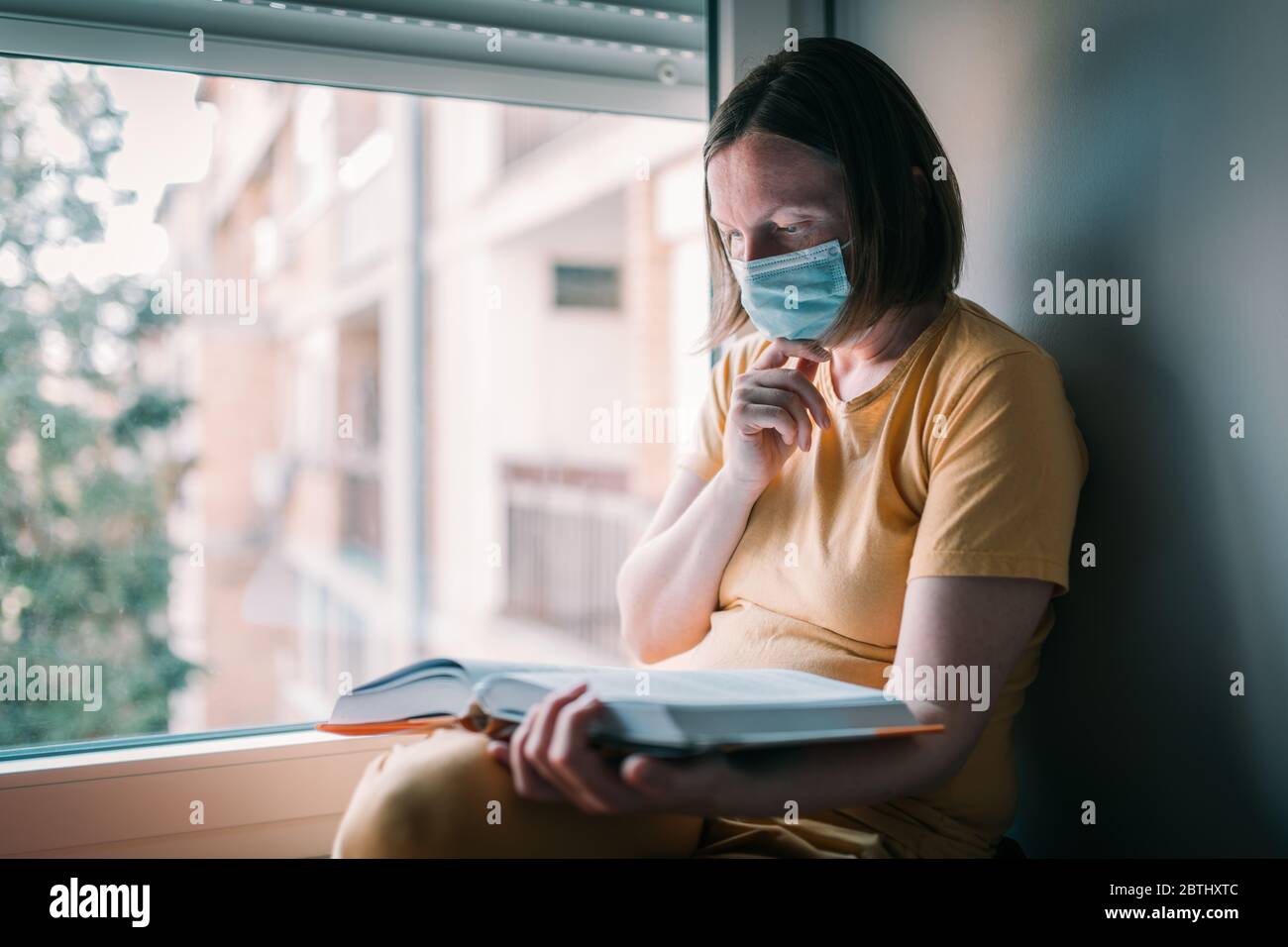 Mujer en el libro de lectura de auto-aislamiento por la ventana. Mujer con mascarilla respiratoria protectora facial en cuarentena domiciliaria. Foto de stock