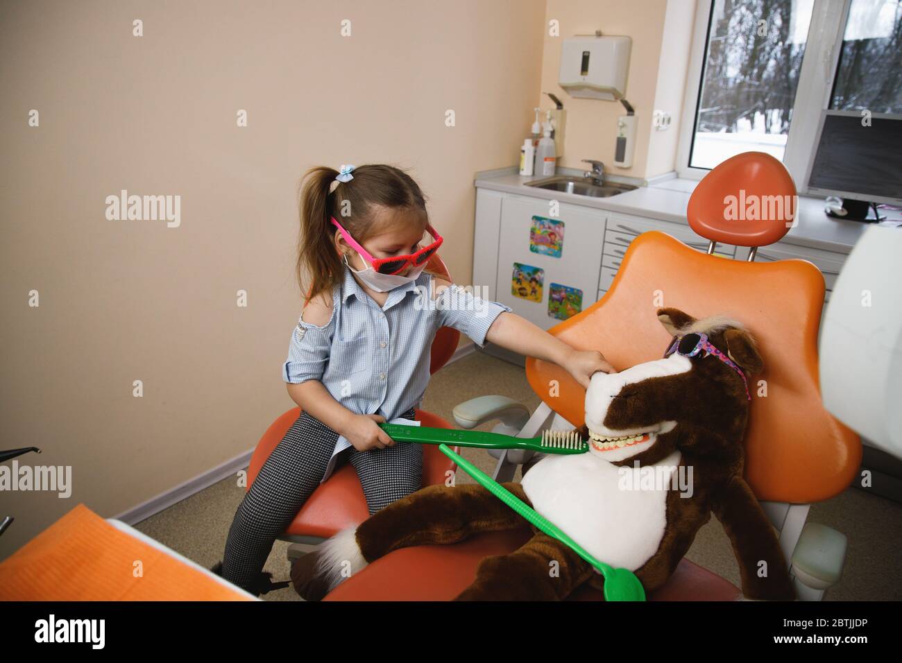 Ángulo alto de niña en máscara médica y gafas de sol cepillando los dientes de caballo de juguete mientras fingen ser dentista Foto de stock