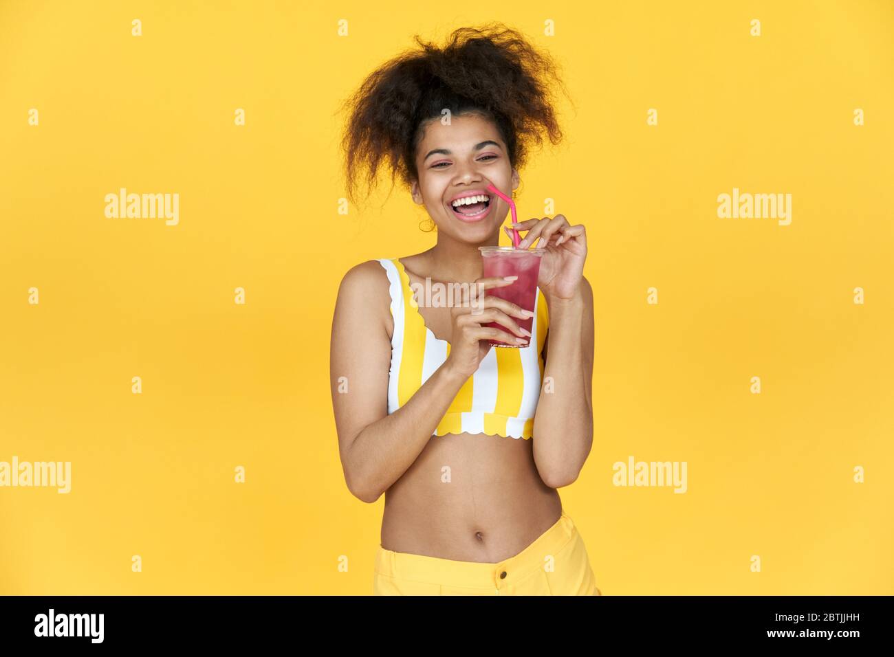 Emocionada niña africana celebrar bebida mirar cámara risa aislada en estudio amarillo. Foto de stock
