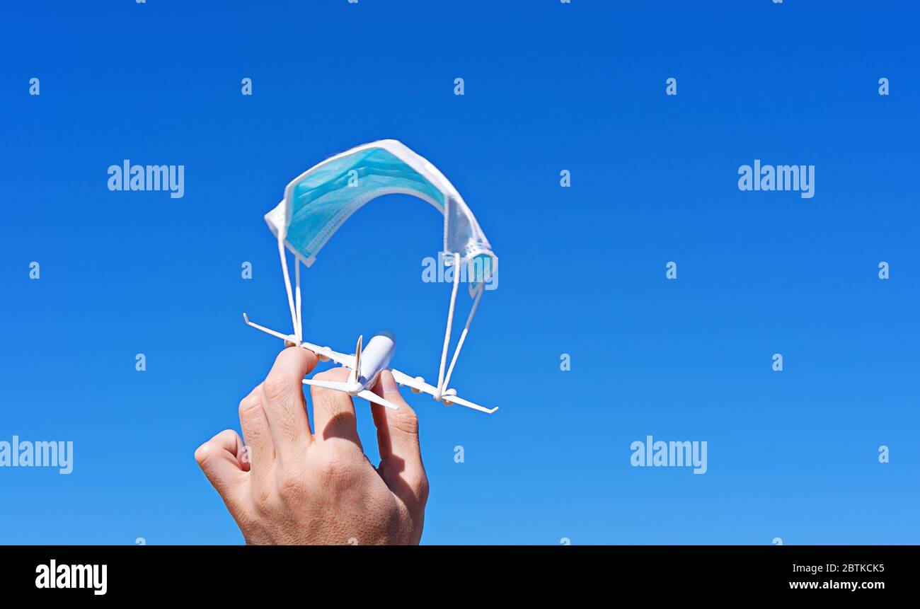 Avión en mano masculina con paracaídas de cara mascarilla quirúrgica contra el cielo azul. Nuevas normas seguras en cabina de aeronaves. Prohibición de vuelos es concepto suspendido, espacio de copia Foto de stock