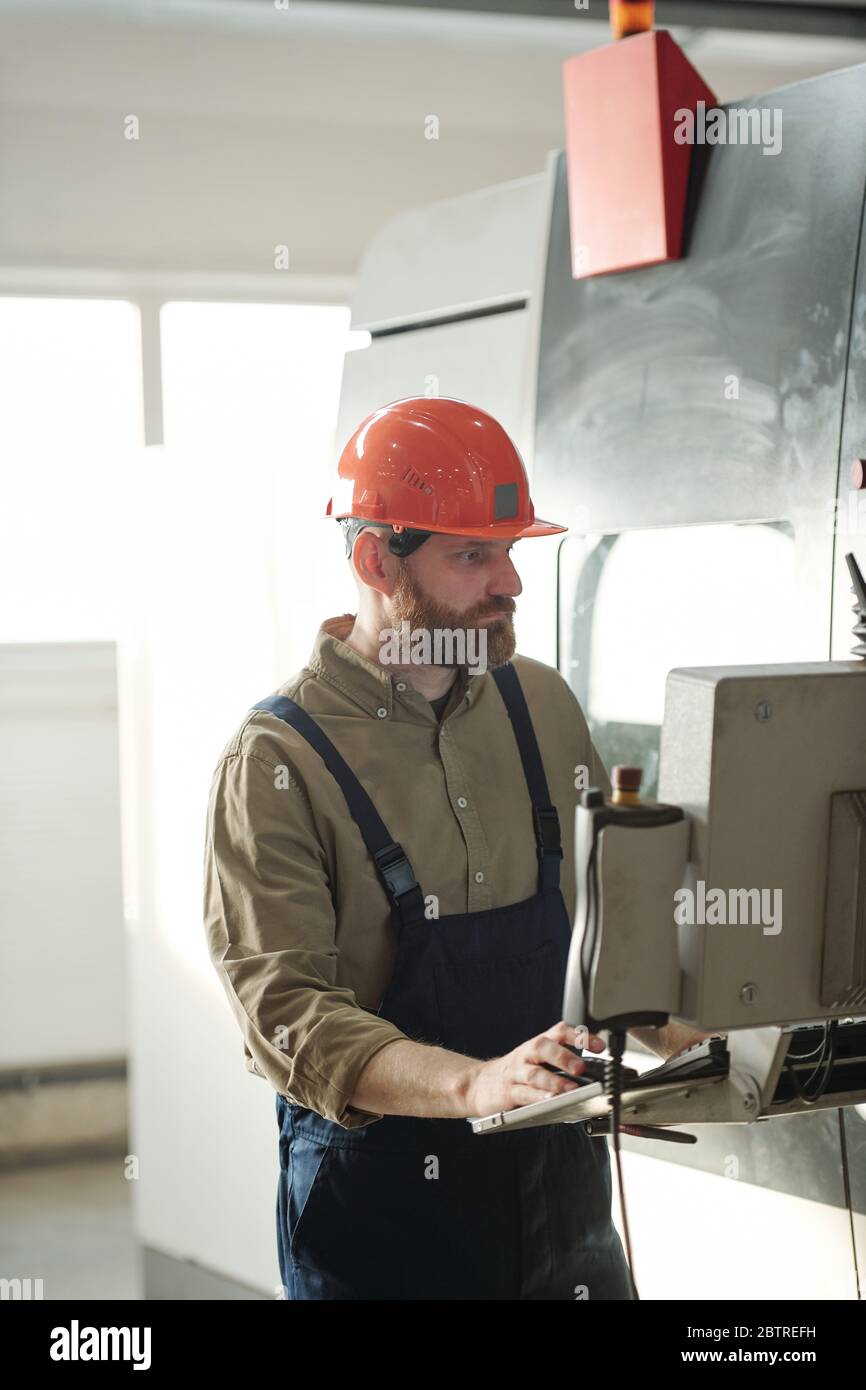 Operador de barba concentrado en casco y overoles elegir configuraciones en  la computadora de la máquina CNC mientras trabaja en la fábrica Fotografía  de stock - Alamy