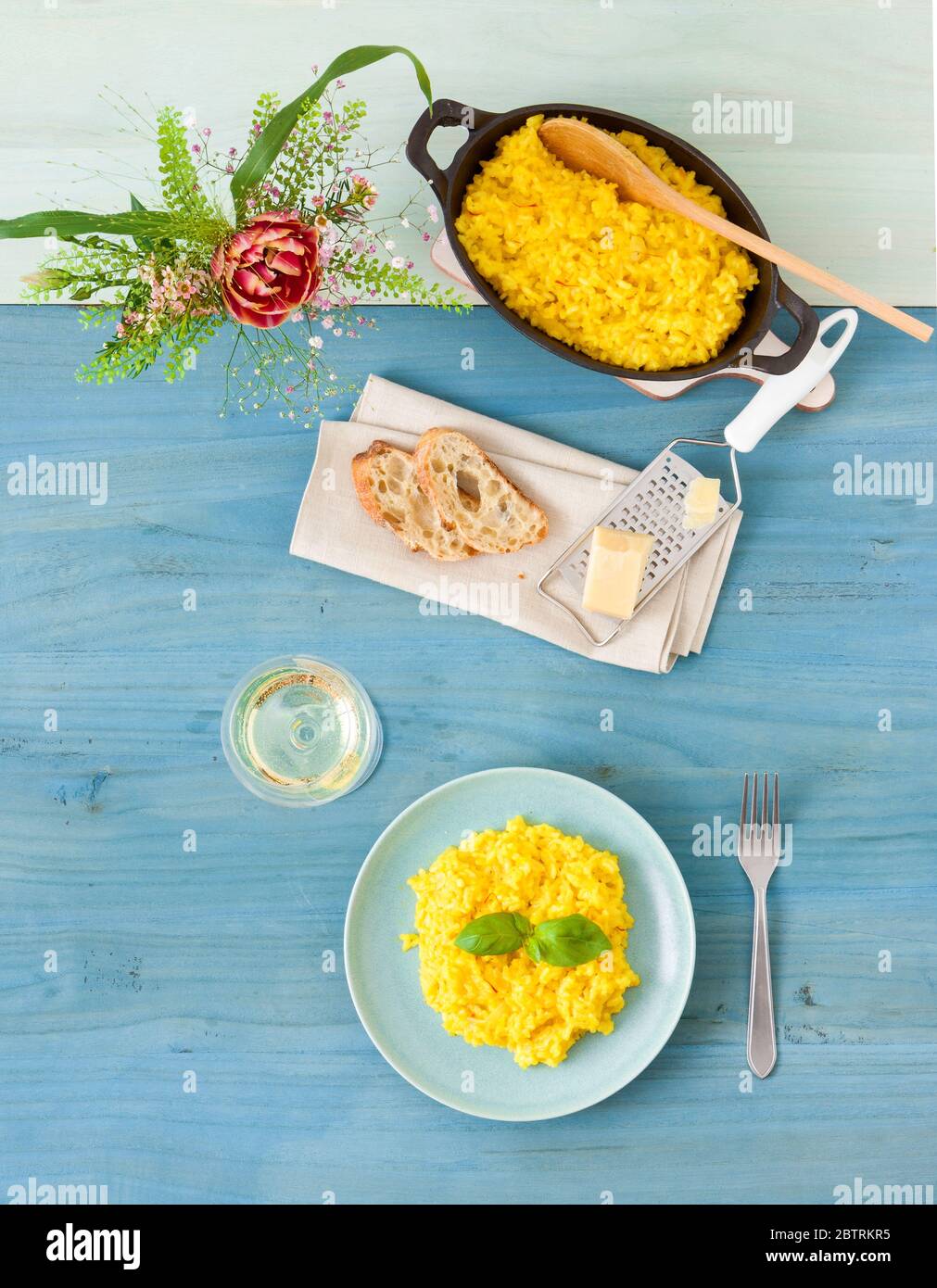 Risotto Milanese, mesa de madera con risotto de azafrán italiano tradicional, vasos y jarra de vino, botella de aceite de oliva, cesta de pan y flores Foto de stock