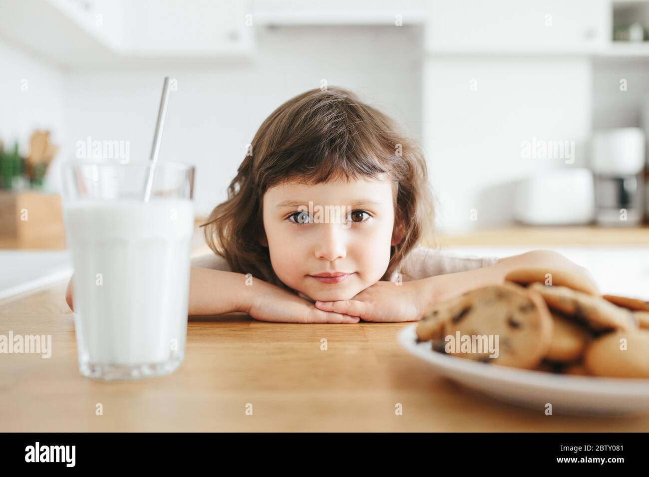 Linda niña pequeña bebiendo leche con paja de acero de vidrio y comiendo galletas sentadas en la mesa de la cocina. Reduzca el uso de plástico en casa con los niños Foto de stock