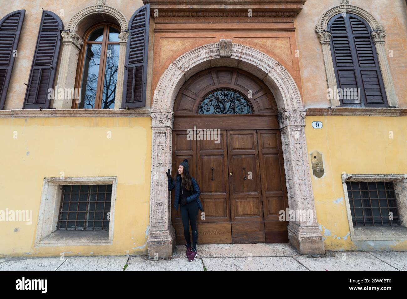 Imagen horizontal de una joven morena posando frente a una enorme puerta de madera en Verona, Italia Foto de stock