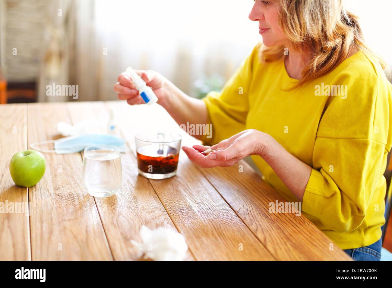 Vista lateral de la cosecha de la mitad de edad femenina tomando remedio de la botella de plástico mientras se sienta en la mesa con una taza de té y máscara protectora en casa Foto de stock