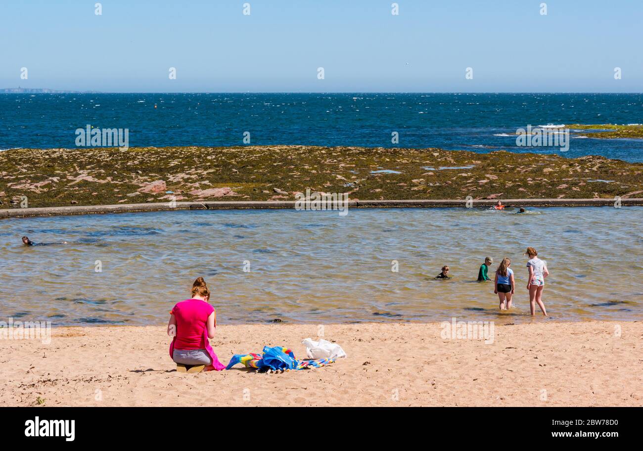 Berwick del Norte, Lothian del este, Escocia, Reino Unido, 30 de mayo de 2020. El tiempo en el Reino Unido: Los que buscan sol en el popular complejo costero en un día soleado, pero muy cálido, hacen que la ciudad vuelva a la normalidad después de un descanso cerrado. Gente en la playa este en la bahía de Milsey. Una mujer se sota en la playa mientras los niños reman en la piscina de agua de marea en el clima cálido Foto de stock