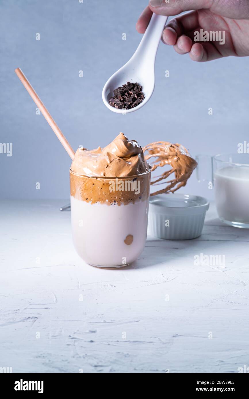 Mano de hombre añadiendo chips de chocolate a un café de Dalgona sobre fondo blanco. Bebida coreana de moda. Imagen vertical. Foto de stock