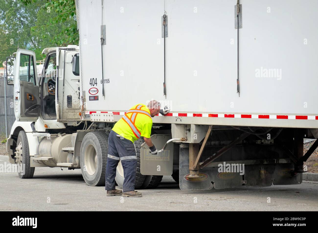 Un conductor de camión que lleva chaleco de seguridad y guantes de trabajo realiza ajustes en un camión semirremolque blanco. Foto de stock. Foto de stock