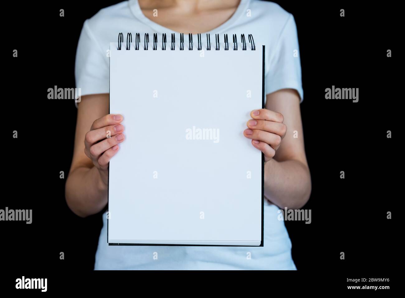 Torso de mujer caucásica fotografiada contra fondo negro, sosteniendo una libreta de notas en espiral en blanco Foto de stock