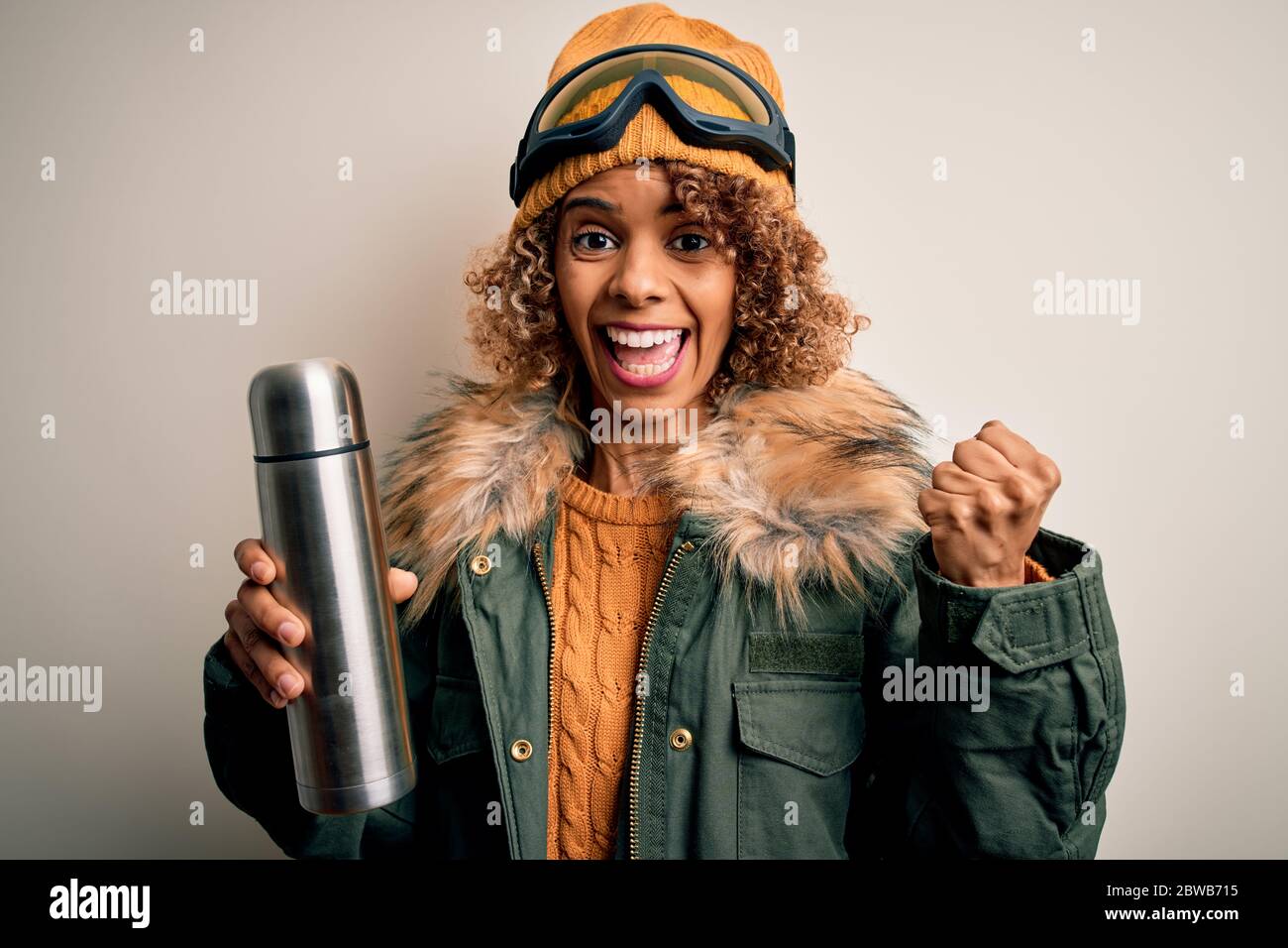 Joven afroamericana esquiadora mujer con gafas de esquí tomando termo con café gritando orgulloso y celebrando la victoria y el éxito muy emocionado, c Foto de stock