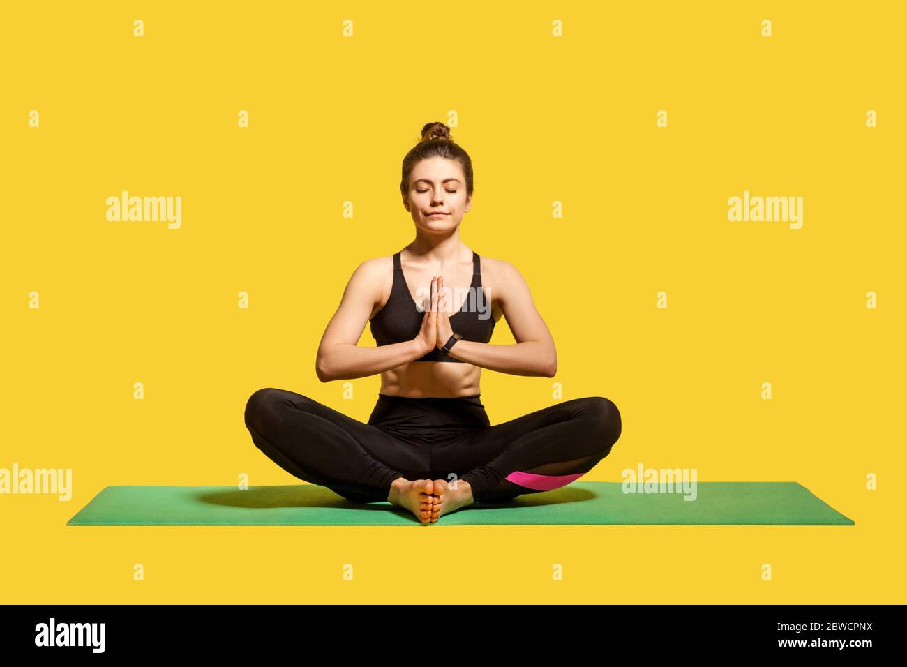 Mujer pacífica concentrada con pelo en el pelo bun apretado sportswear sentado en la estera practicar yoga, sosteniendo las manos en el gesto namaste y meditando, relaxin Foto de stock