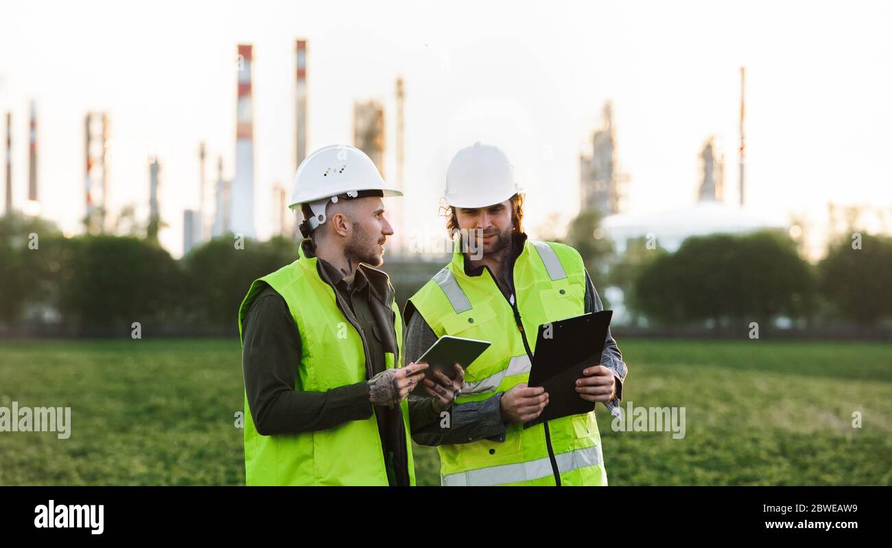 Dos jóvenes ingenieros con tabletas de pie en el exterior por la refinería de petróleo, discutiendo cuestiones. Foto de stock