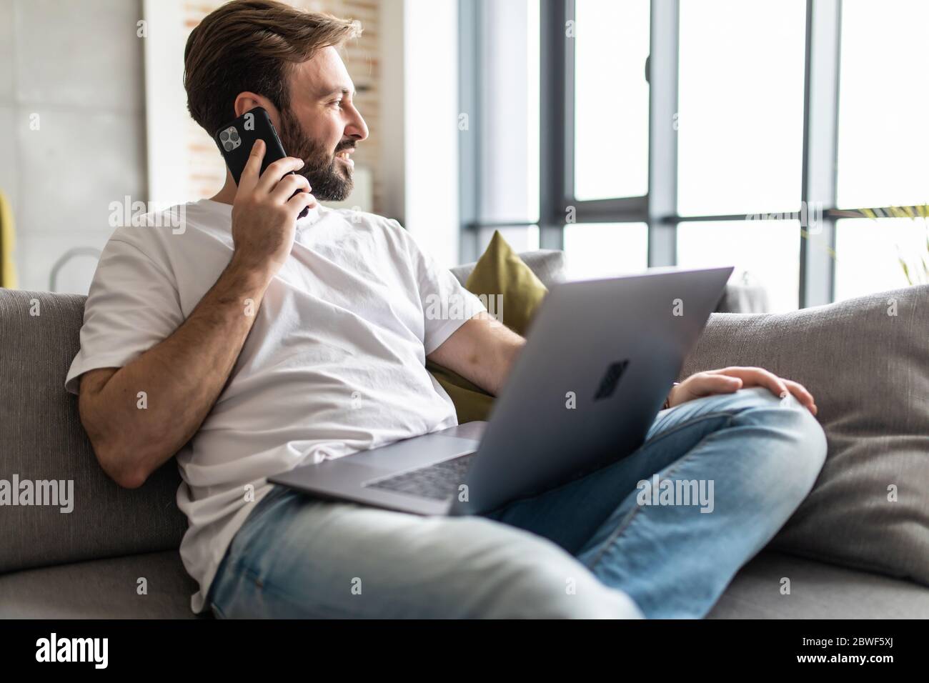 Joven usando un ordenador portátil mientras habla por teléfono sentado en el sofá Foto de stock