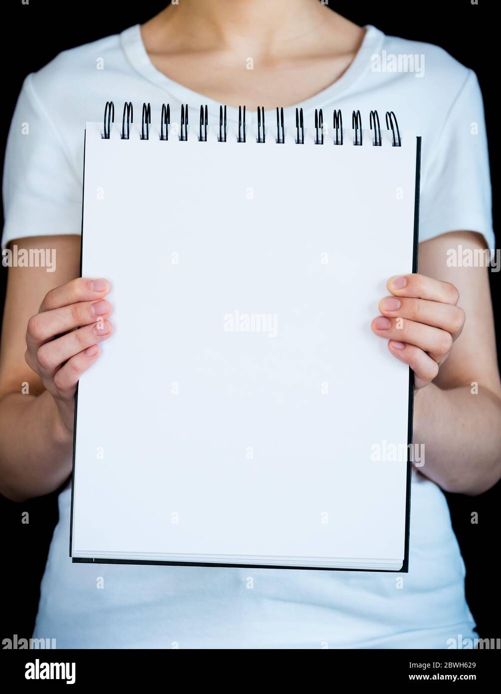 Primer plano de la página en blanco de bloc de notas sostenido por la mujer con camiseta blanca contra fondo negro Foto de stock