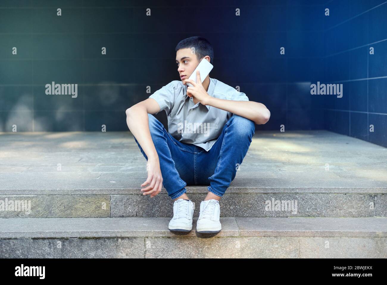 Un joven adolescente charlando en su teléfono móvil al aire libre sentado en los escalones de la ciudad mientras mira lejos al lado escuchando la conversación con un sin embargo Foto de stock